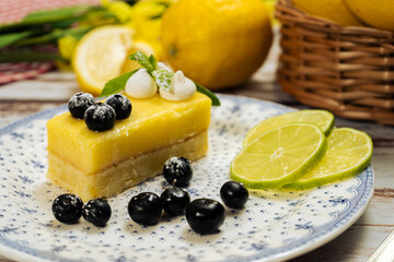 Slice of homemade lemon curd cake in the shape of an ingot or brick decorated with some mint leaves, blueberries and some lemons in the background on an old wooden table.