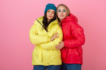 two attractive stylish women posing on pink background in colorful winter down jacket of red and yellow color, warm clothes fashion trend