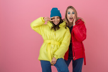 two attractive girl friends active women posing on pink background in colorful winter down jacket of bright red and yellow color having fun together, warm coat fashion trend, sincerely laughing