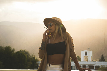 Blond girl with glasses and hat smoking on the terrace of a hotel as sunset falls