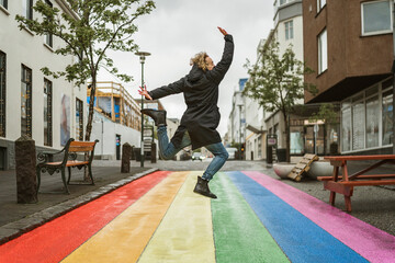 Anonymous boy jumping for joy in the streets of Reykjavik, Iceland on a roadway painted in the colours of the LGTBI+ rainbow flag. Celebrating sexual equality. - obrazy, fototapety, plakaty