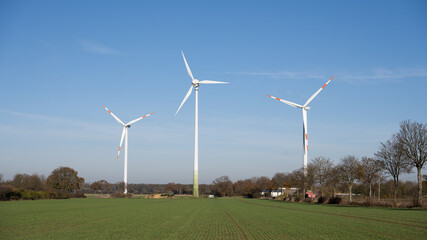 Fototapeta premium Three red and white wind turbines for power generation in Germany with a blue sky in the background and a green field in the foreground