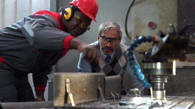 African American CNC Machine Operator Talking To Caucasian Quality Control Inspector Next To Milling Machine In Action - CNC Milling Machine Tool Cutting Steel - Rack Focus