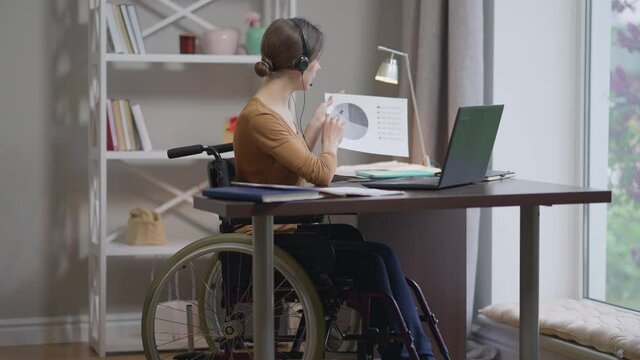 Portrait of confident Caucasian young woman in wheelchair talking at laptop video chat pointing at graph. Beautiful slim freelancer working messaging online from home office. Remote working freelance