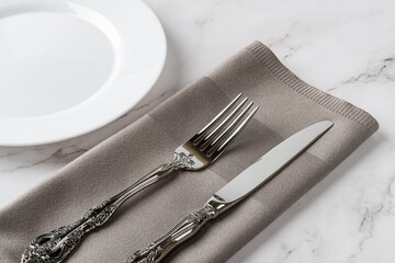 Elegant vintage fork and table knife on a gray dinner napkin and white porcelain plate over marble tabletop. Crockery and cutlery for set table. Empty clean ceramic plate for food design mockup.