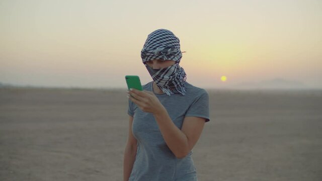 Woman in checkered keffiyeh standing in desert using mobile phone. Caucasian female tourist taking pictures of desert during sunrise.