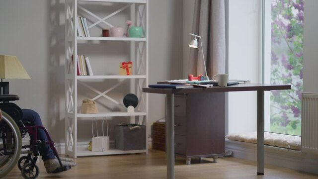 Wide shot home office with disabled young woman rolling wheelchair in slow motion to table opening laptop. Portrait of serious professional Caucasian expert freelancer indoors working online