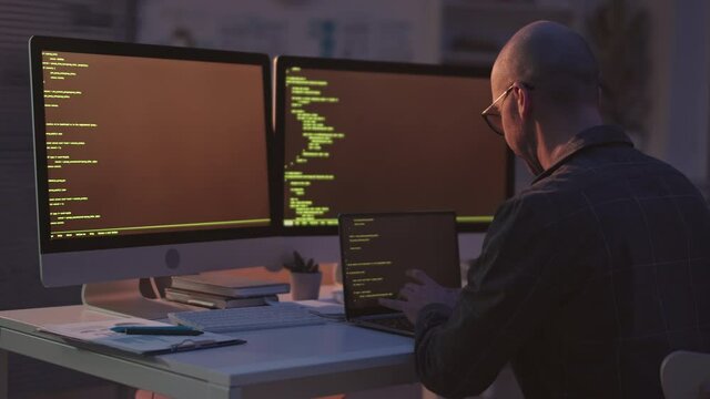 Rear-view medium slowmo shot of bald Caucasian programmer writing program codes on laptop sitting at desk with two monitors in office at night
