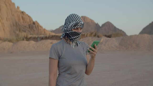 30s female tourist in checkered keffiyeh talking on mobile phone standing in the desert. Woman using cell in desert and bedouin with camel walking by.
