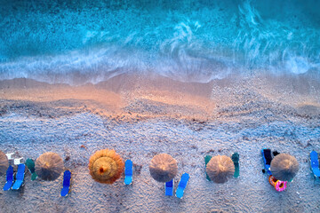 Aerial, vertical view of a gravel beach with colorful sunbeds and reed umbrellas, deep azure sea. ...