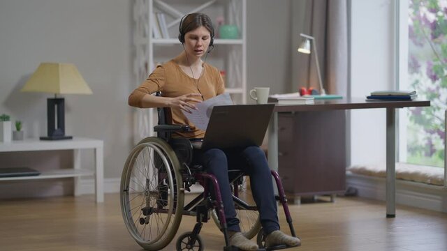 Disabled expert young woman in headphones talking showing graph at laptop web chat persuading colleagues in idea profitability. Wide shot portrait professional intelligent Caucasian freelancer indoors