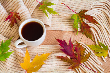 A cup of coffee wrapped in a warm knitted scarf and fallen autumn leaves on a wooden table. Autumn home coziness concept