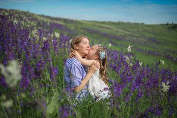 mom and daughter are sitting on the grass in a beautiful field with purple flowers on a sunny day. The girl hugs and kisses her mother.