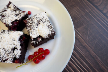 close by on a white plate on a wooden table are three pieces of gluten-free brownies with red currants sprinkled with powdered sugar. gluten- and lactose-free cakes. homemade cakes with a diet