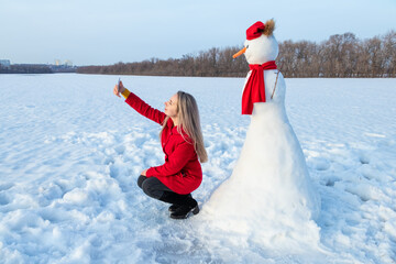 Obraz premium Attractive woman in red coat taking selfie with snowman using smartphone