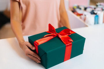 Close-up of unrecognizable female hands holding wrapped Christmas gift box tied red ribbon and decorated beautiful bow, closeup. Xmas or New year gift in stylish wrapping paper with red ribbon.