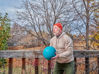 senior male (on late 60s) is exercising with a heavy slam ball in his backyard, chilly afternoon in...