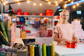 Smiling young woman holding and using golden ribbon to tie bow for red wrapped gift box for Christmas present at counter of holiday shop, looking at camera, blurred background of bright lighting