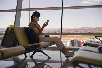 Woman in face mask waiting boarding in big empty airport. Female traveler look at phone, search for boarding pass or flight cancelation information.