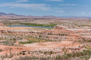 Lake Mead, Nevada, USA - May 22, 2011: Red and gray rock landscape at edge of blue water lake under blue cloudscape with scarce sprinkled green vegetation.