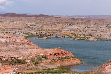 Fototapeta na wymiar Lake Mead, Nevada, USA - May 22, 2011: Wide landscape with blue water lake in center and dry beige-red rocky shores and hills under light blue sky. Small fishing boat in center.