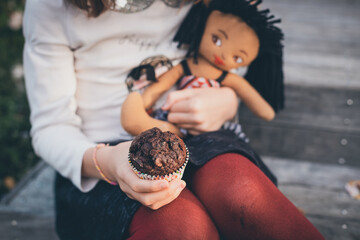 kid holding a chocolate banana muffin next to a doll - obrazy, fototapety, plakaty