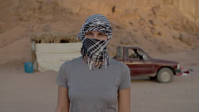 Portrait of woman wearing black and white checkered keffiyeh standing in the desert looking at camera. Caucasian female tourist taking off headdress and smiling at camera.