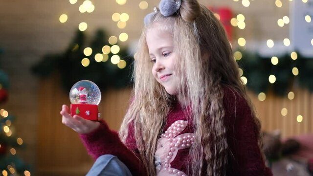 The girl holds a souvenir in the form of a glass ball with snow in her hand and shakes it. Festive atmosphere before Christmas and New Year