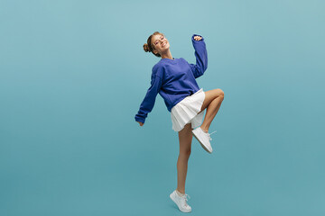 Full length funny young european girl dancing on blue background in studio. Fair-haired happy teenager smiles broadly with two tufts on her head, wearing sweatshirt and skirt. Holiday life concept