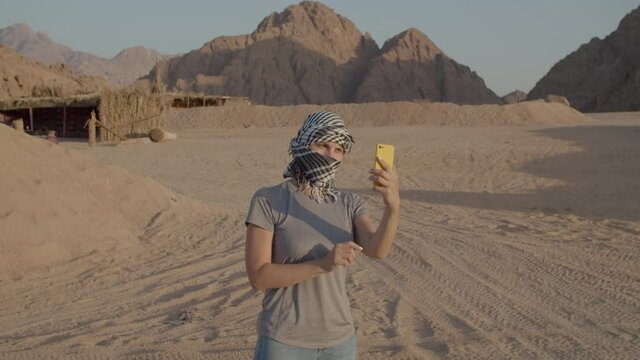 Woman in checkered keffiyeh trying to find mobile network using cell phone standing in desert. Person searching for mobile signal in desert. No cell operator signal.