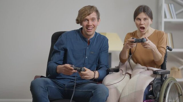 Joyful laughing young couple playing video game with joysticks having fun on weekend indoors. Portrait of positive disabled Caucasian girlfriend and loving boyfriend enjoying gaming in slow motion