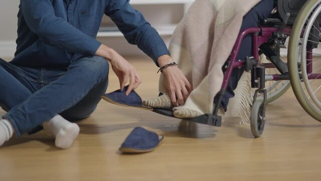 Unrecognizable loving man putting slippers on legs of disabled woman in wheelchair in slow motion. Caucasian husband boyfriend talking care of handicapped wife girlfriend indoors at home. Love support