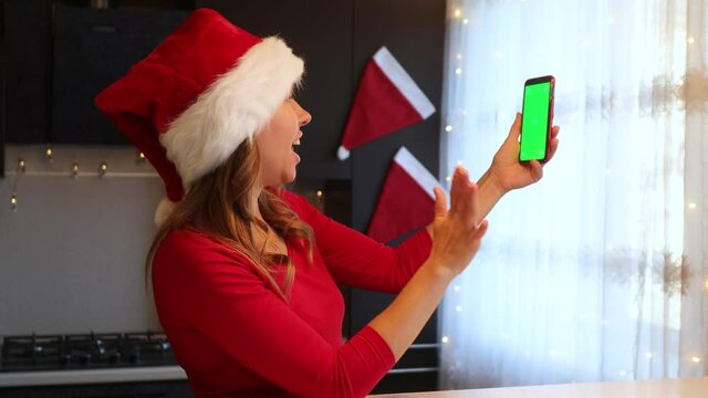 A girl in a New Year's red suit in the kitchen leafs through a smartphone with a green screen with her finger
