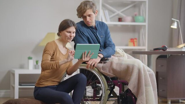 Positive loving young woman supporting man in wheelchair surfing Internet on tablet talking smiling. Happy Caucasian girlfriend and disabled boyfriend indoors at home. Endorsement and care