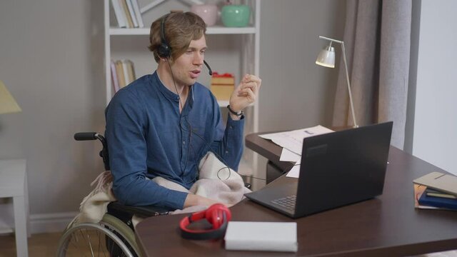 Positive confident male disabled freelancer conferencing online in slow motion smiling. Portrait of happy Caucasian young paraplegic man talking at laptop video chat sitting in wheelchair indoors