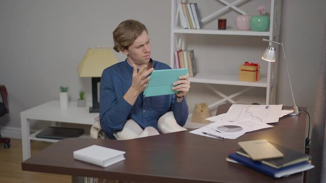 Intelligent disabled young man in wheelchair messaging online on tablet sitting in home office indoors. Portrait of confident Caucasian positive freelancer texting e-mail smiling in slow motion
