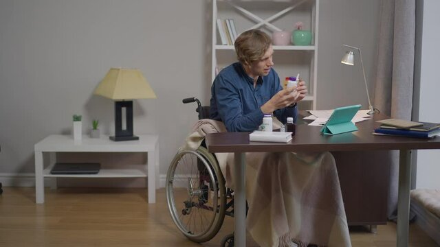 Wide shot young paraplegic man in wheelchair consulting doctor online talking showing pills at tablet video chat. Portrait of Caucasian disabled guy asking advice in slow motion. Modern technologies