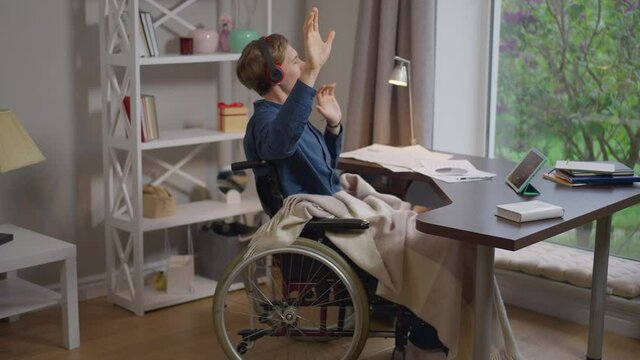 Happy young disabled man in wheelchair listening to music in headphones. Wide shot portrait of Caucasian paralyzed guy in home office on break enjoying hobby in slow motion. Lifestyle and joy