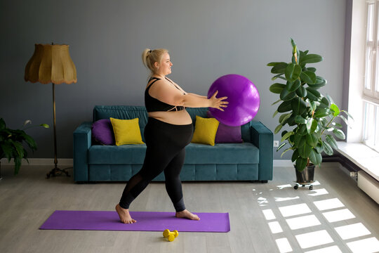 A fat, obese pregnant lady is doing Pilates at home, lifting a fit ball above herself. Home workouts in Interior room with a large window, sofa and yellow-purple pillows. Weight loss in quarantine  - Powered by Adobe