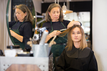 Young brown haired woman sitting in chair in hairdressing studio while elderly female stylist making hairstyle, cutting hair.