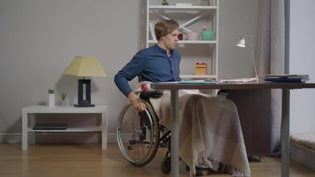 Serious talented disabled architect rolling wheelchair from table in home office with rolled blueprint on knees. Portrait of professional expert paralyzed Caucasian man indoors. Slow motion