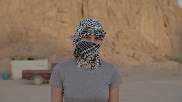 30s woman wearing black and white checkered keffiyeh walking in the desert looking at camera. Caucasian female tourist taking off headdress and smiling at camera.