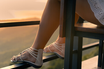 Close up of the legs of a young woman in white dress and sandals sitting on a viewpoint at sunset. Mountains and sea in the background 