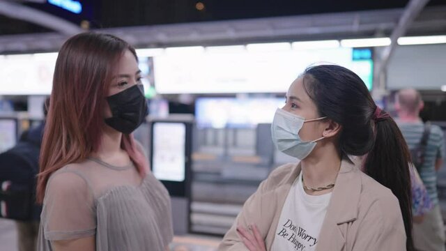 Two young adult asian women wear protective facemask talking to each other on the skytrain platform while que up waiting for the train, new normal life after pandemic, people adapt to new lifestyle