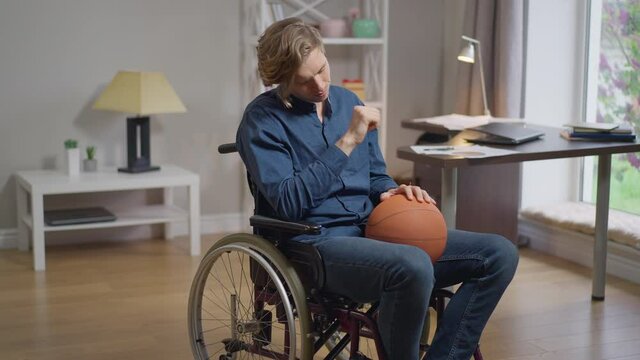 Live camera zoom in desperate Caucasian disabled man in wheelchair with basketball ball. Portrait of hopeless paraplegic sportsman thinking on future sport career at home indoors. Slow motion