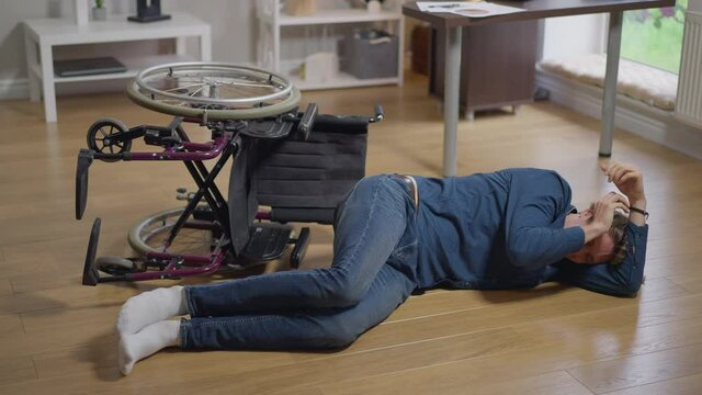 Wide shot crying disabled man lying on floor with turned over wheelchair. Desperate lonely frustrated Caucasian paralyzed guy alone at home indoors. Loneliness and disability concept