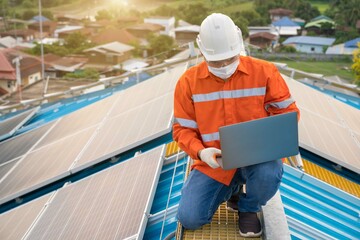 Industrial technicians wearing helmets and COVID-19 protective masks use laptop computers to monitor solar roof maintenance. Periodic checks are performed to preserve and prolong solar panels.