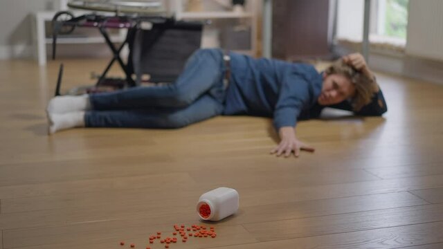 Wide shot blurred disabled young man lying on floor fallen from wheelchair reaching to pills. Paralyzed Caucasian stressed guy making effort stretching to medications in slow motion