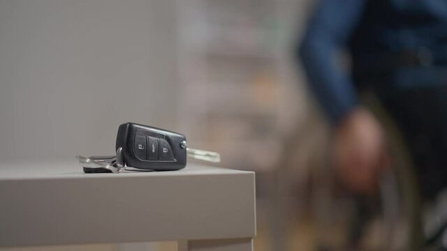 Close-up car key indoors with blurred disabled paralyzed man rolling wheelchair at background leaving. Unrecognizable injured Caucasian young guy after auto accident at home indoors