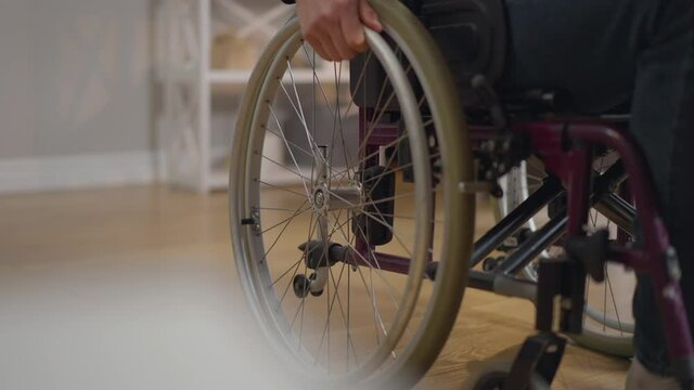 Unrecognizable disabled man rolling wheelchair in slow motion leaving indoors. Young Caucasian paralyzed guy at home in living room. Illness and disability concept